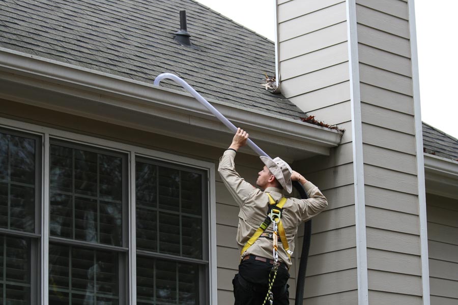 A technician cleans a gutter on the roof above him using a curved vacuum attachment.