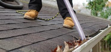 The lower legs and boots of a franchisee are visible on a rooftop as the franchisee vacuums leaves and other debris out of a client’s gutter.