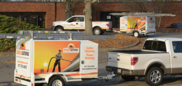 Two Outback GutterVac pickup trucks with equipment trailers sit parked outside a commercial building, with leaves littering the ground around them.
