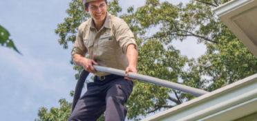 A smiling franchise owner cleans gutters with a vacuum on top of a client’s roof.