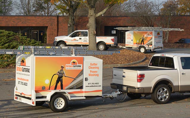 Two Outback GutterVac trucks with equipment trailers are parked outside a leafy office park.